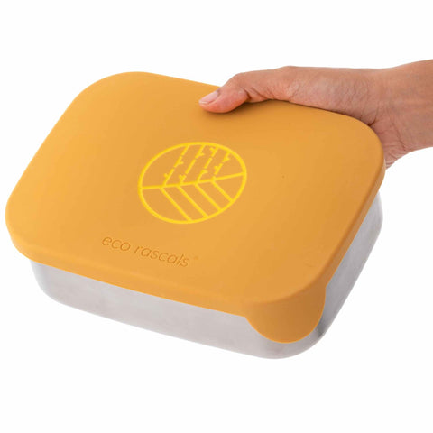 Image shows the classic Mustard eco rascals lunch box bento style with removable dividers being held closed by an adult hand.